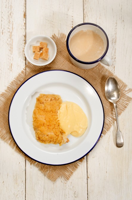 british apple crumble with custard on enamel plate, mug milk coffee and bowl with brown sugar cubes